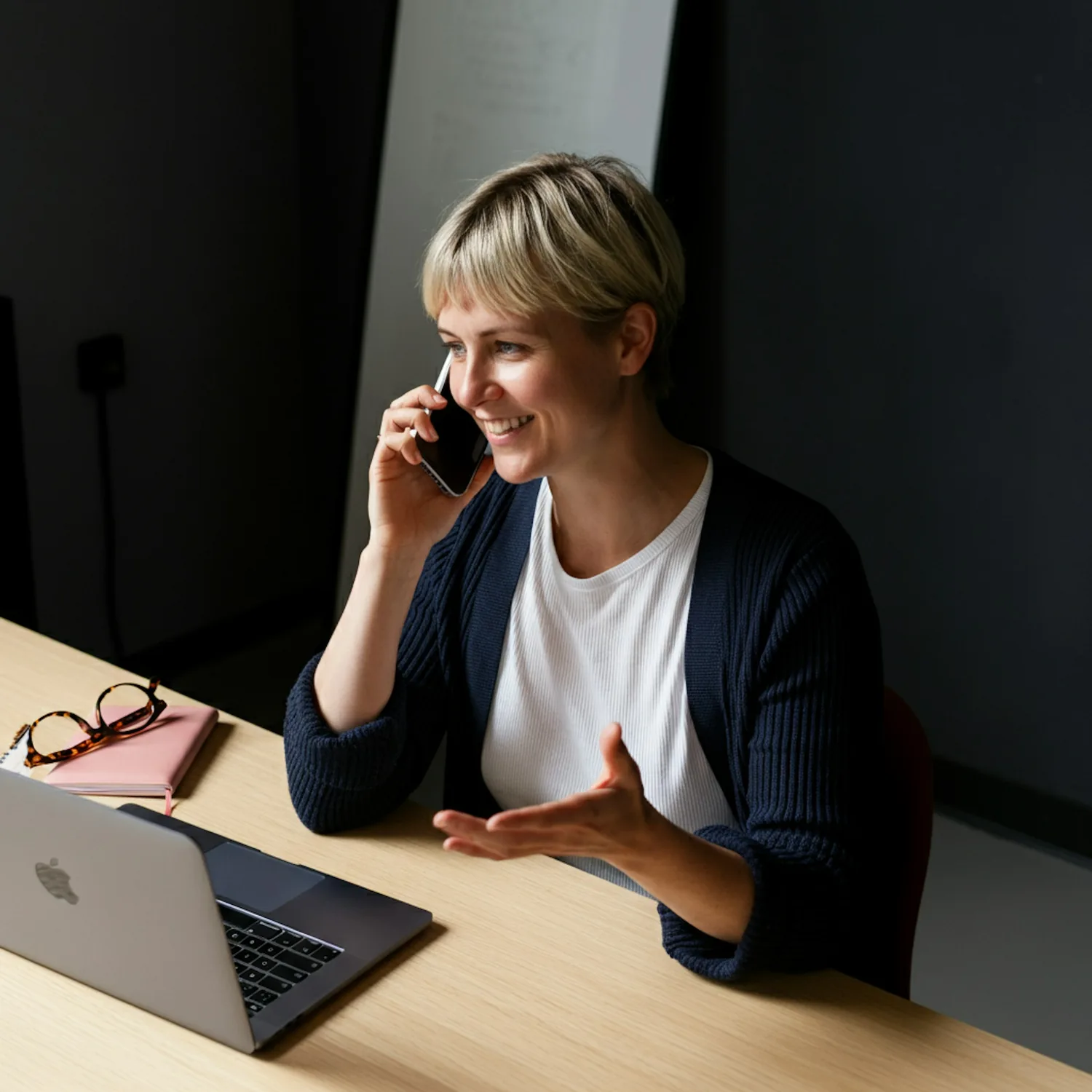 A woman smiling mid-call at her desk, a laptop open beside her
