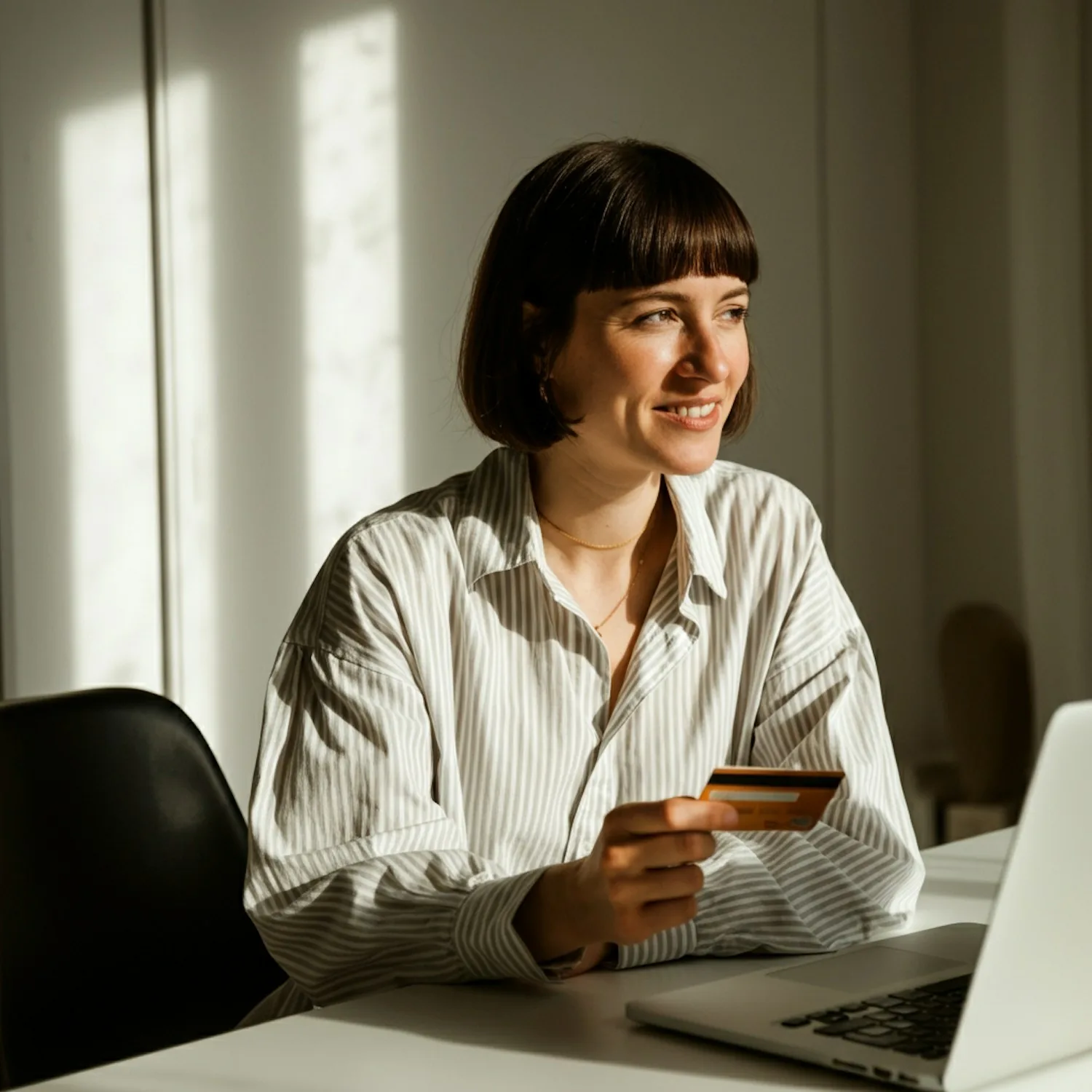 A shopper at a desk in warm sunlight, holding a bank card beside an open laptop